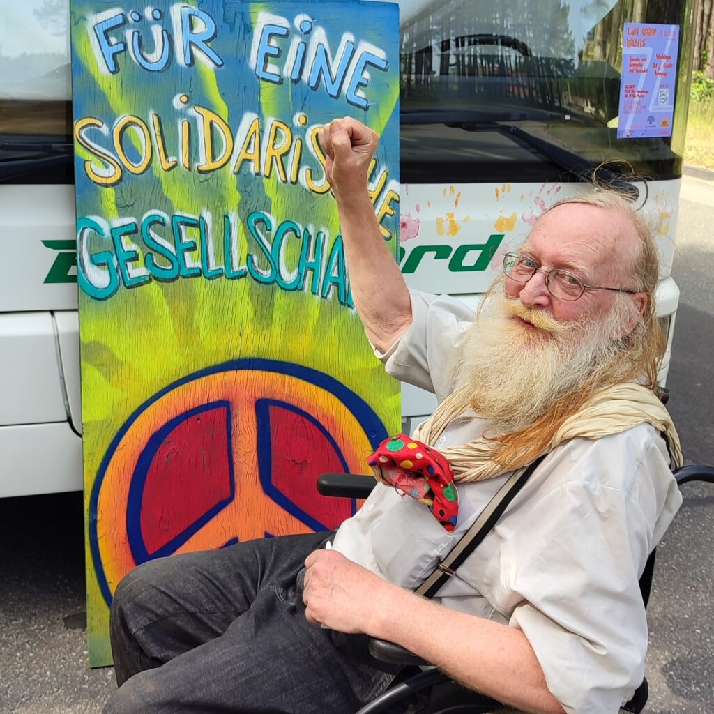 Das Bild zum Thema Solidarische Ökonomie zeigt einen älteren Mann mit langem Bart und erhobener Faust im Rollstuhl sitzend vor einem Plakat mit der Aufschrift "Für eine Solidarische Gesellschaft." Das Plakat stammt von einem Graffiti-Workshop von Einsweiter.