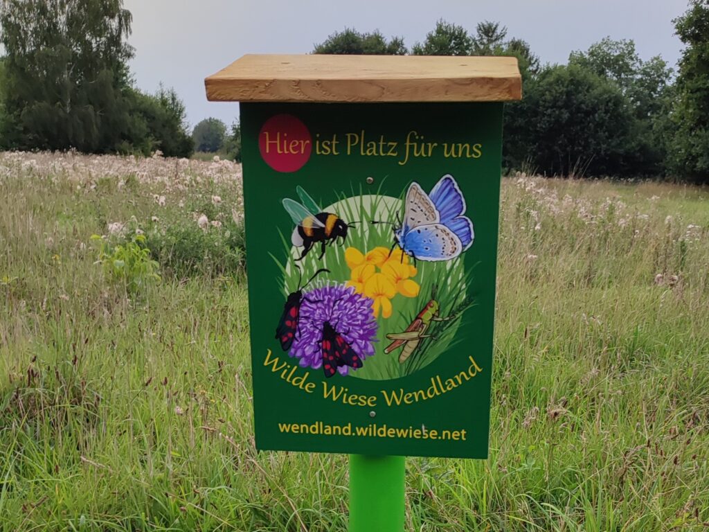 Vor dem Naturgrundstück steht das Hinweisschild der "WILDE WIESE WENDLAND" es zeigt Insekten (Hummel, Schmetterling, Falter und Heuschrecke) und Blumen. Darüber die Aufschrift "Hier ist Platz für uns" 