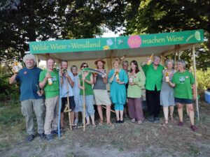 Das Bild zeigt 12 Menschen, die im Freien vor einem Stand stehen. Der Stand trägt den Namen "WILDE WIESE WENDLAND".  Es sind die Mitglieder der Aktions-Gruppe "Wilde Wiese". 