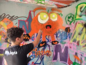 Ein Bild von unsere Graffiti-Workshop auf dem Einsweiter-Gelände. Ein Junge malt ein kreisrundes, orangefarbenes Wesen mit großen gelben Augen. 