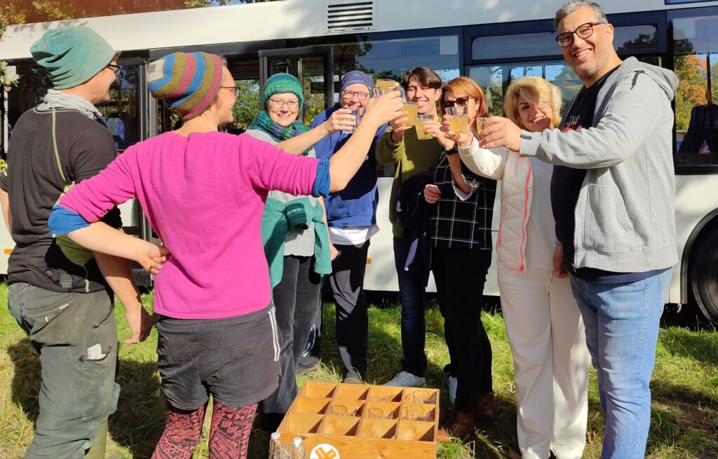 Das Bild zum Thema Mitmachen zeigt eine Gruppe Helfer*innen, die auf dem Einsweiter-Gelände vor dem Bus steht und mit Getränken anstößt. Sie machen gerade Pause von der gemeinsamen Wiesen-Pflege.